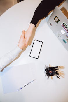 Two individuals shaking hands across a desk with a smartphone and laptop in a modern office setting.