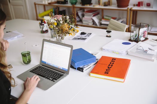 Woman works on a laptop in a stylish office, surrounded by books and documents.